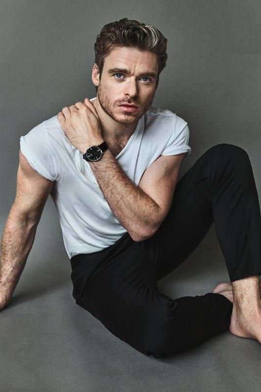 Indoor portrait of a man in a white t-shirt and black pants sitting on a gray background, looking at the camera with a neutral expression.