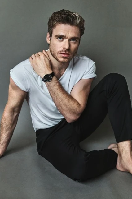 Indoor portrait of a man in a white t-shirt and black pants sitting on a gray background, looking at the camera with a neutral expression.