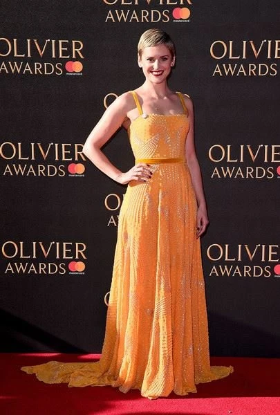 A woman with short blonde hair poses on a red carpet wearing a long, yellow dress at the Olivier Awards, smiling at the camera.