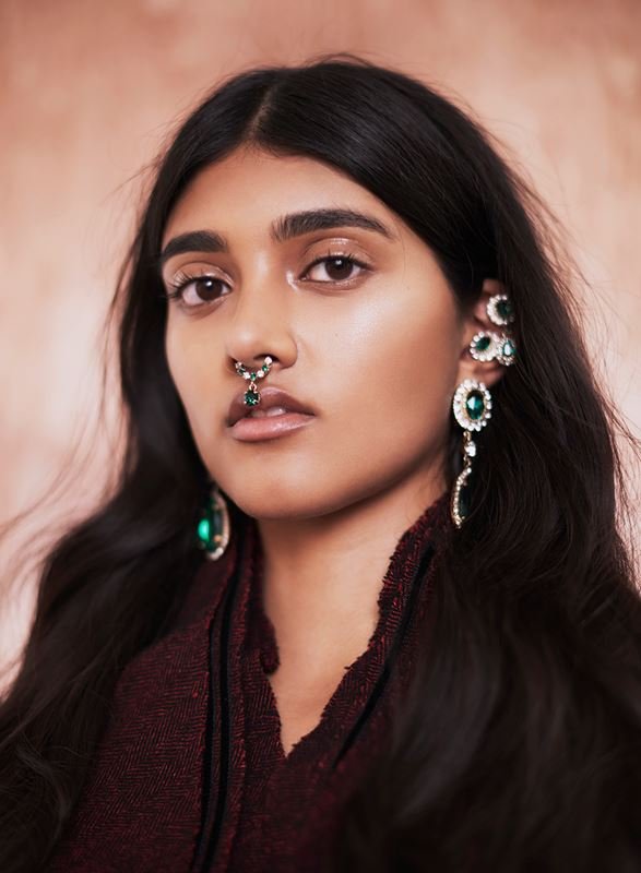 Close-up portrait of a woman with dark hair, wearing statement jewelry, against a textured pink background in a studio setting.