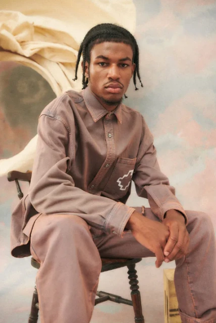 Young adult man in a brown long-sleeve shirt and matching pants sits on a wooden chair against a painted backdrop.