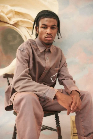 Young adult man in a brown long-sleeve shirt and matching pants sits on a wooden chair against a painted backdrop.