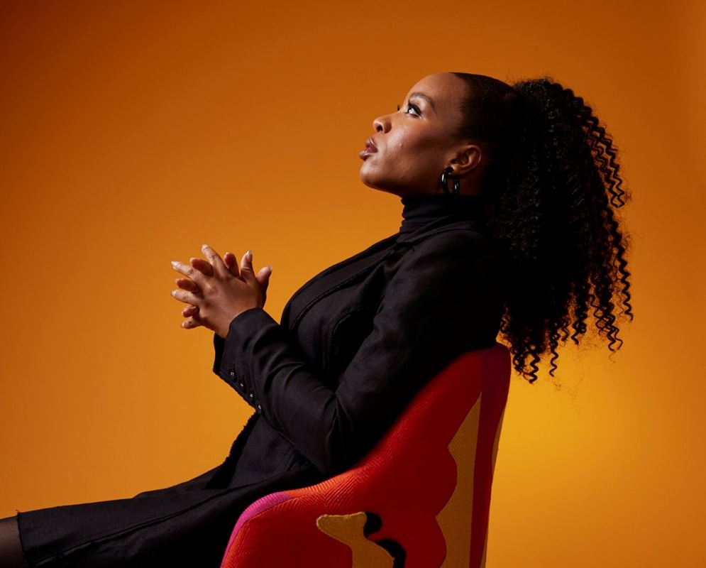Profile of a woman with curly hair, wearing black blazer, sitting in a chair against an orange backdrop, looking upward with hands clasped.