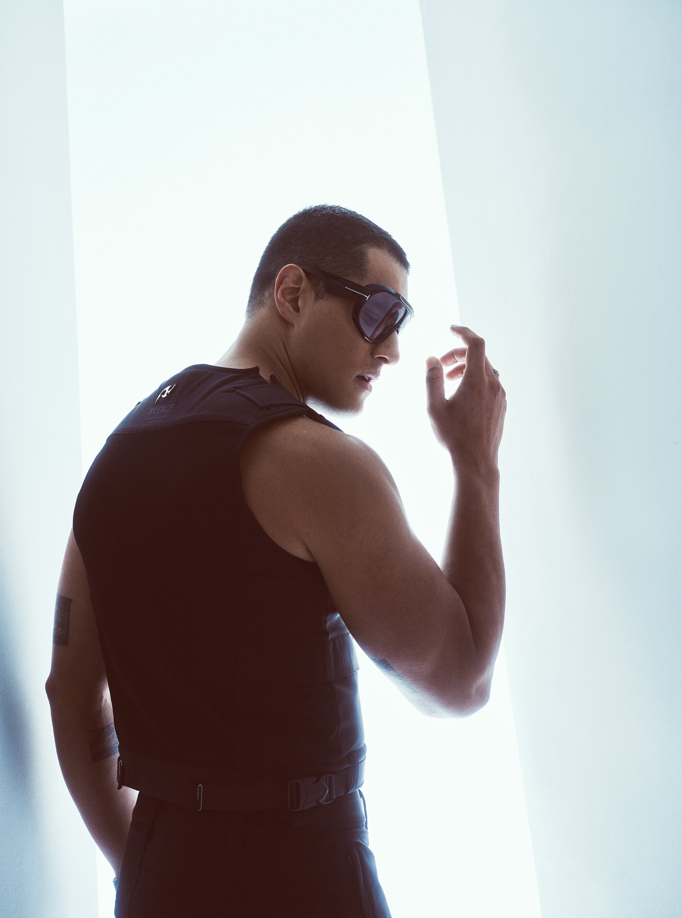 A man poses in dark vest and sunglasses, looking over his shoulder in bright studio lighting, creating a modern fashion photograph.