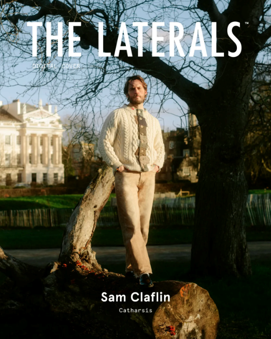 Sam Claflin poses outdoors in a sweater and pants, standing on a fallen tree in front of a building and a large tree. Magazine cover.