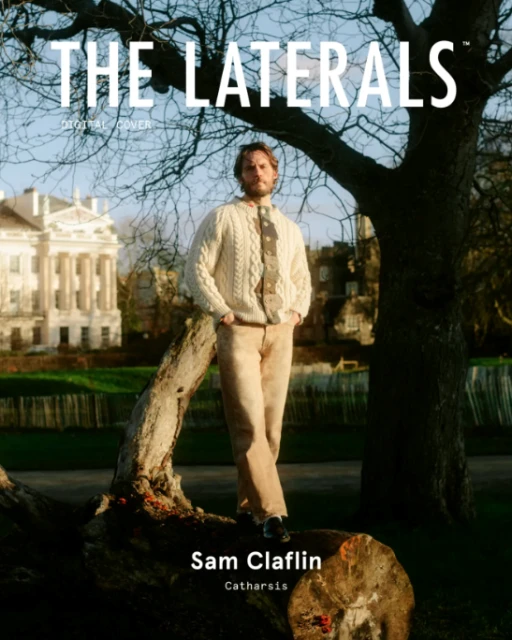 Sam Claflin poses outdoors in a sweater and pants, standing on a fallen tree in front of a building and a large tree. Magazine cover.