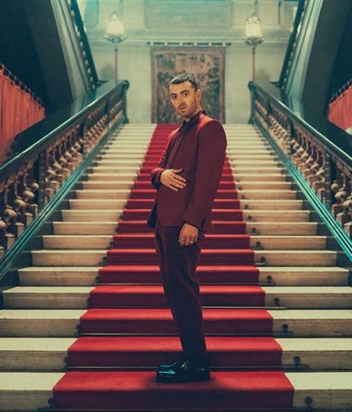 Sam Smith poses on a grand staircase in a red suit, under chandeliers, creating a visually striking and elegant scene.