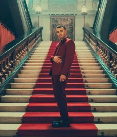 Sam Smith poses on a grand staircase in a red suit, under chandeliers, creating a visually striking and elegant scene.