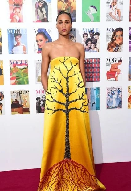 Adult female wearing a yellow strapless gown featuring a tree design, posing against a backdrop of Vogue magazine covers.