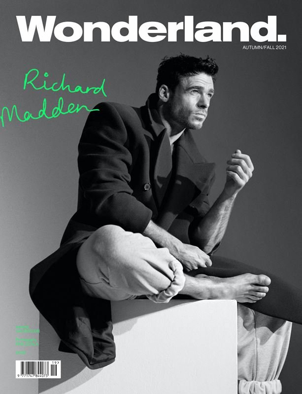 Magazine cover featuring Richard Madden in a dark coat and light pants sitting on a white block against a grey background.