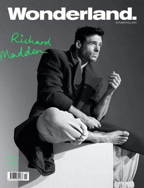 Magazine cover featuring Richard Madden in a dark coat and light pants sitting on a white block against a grey background.