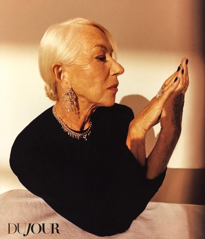 Mature woman with short white hair and diamond jewelry poses with hands near her face in front of a tan background in a studio setting.