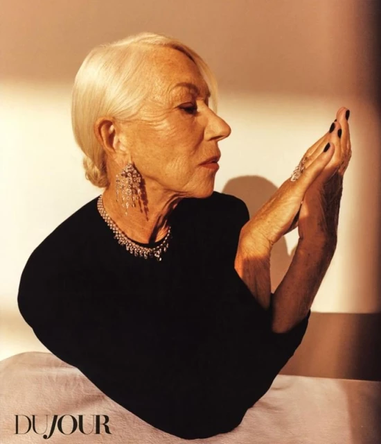 Mature woman with short white hair and diamond jewelry poses with hands near her face in front of a tan background in a studio setting.