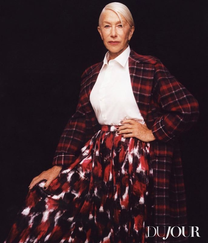 Helen Mirren poses in a red plaid blazer and long skirt with a white shirt against a dark backdrop for a fashion shot.