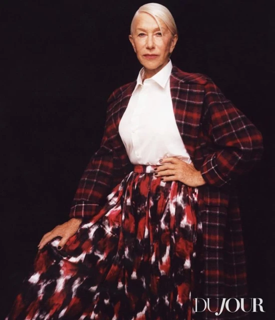 Helen Mirren poses in a red plaid blazer and long skirt with a white shirt against a dark backdrop for a fashion shot.