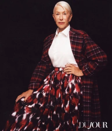 Helen Mirren poses in a red plaid blazer and long skirt with a white shirt against a dark backdrop for a fashion shot.