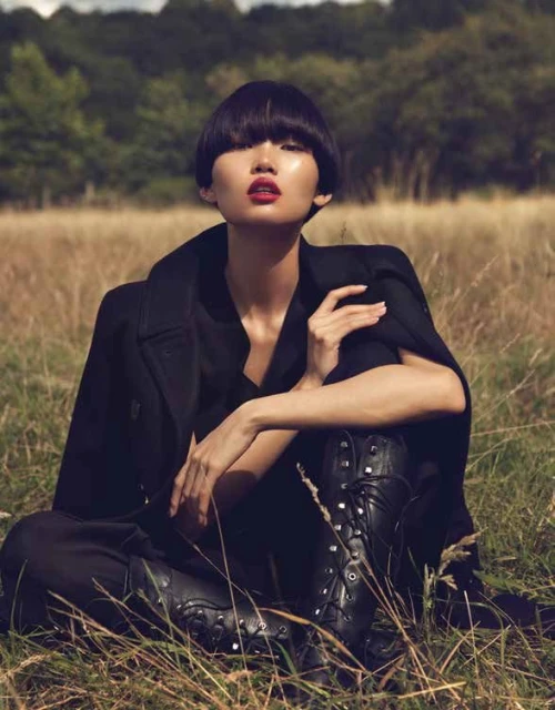 An Asian woman with a blunt hairstyle sits in a field of dry grass wearing a black coat and boots, looking towards the camera.