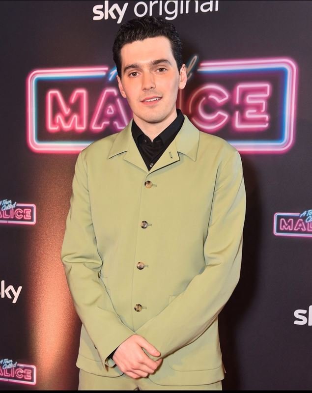 Portrait of an adult man in a suit jacket, black collared shirt, standing in front of a neon sign for a televised show.
