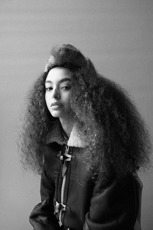 Monochrome portrait of a woman with curly hair and a fur hat wearing a shearling jacket in a studio with a gray background.