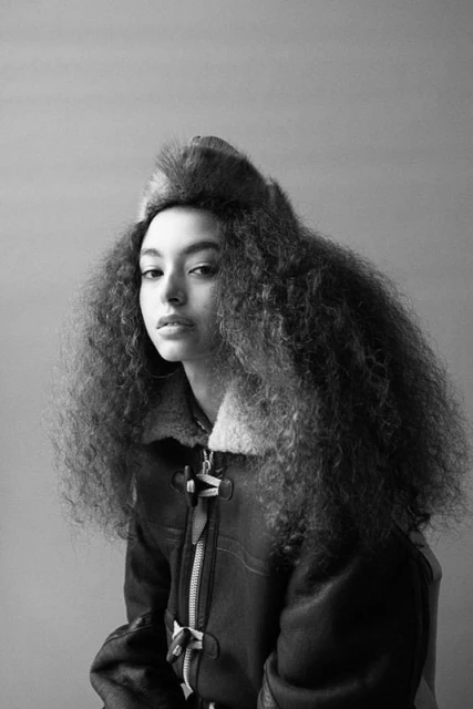 Monochrome portrait of a woman with curly hair and a fur hat wearing a shearling jacket in a studio with a gray background.