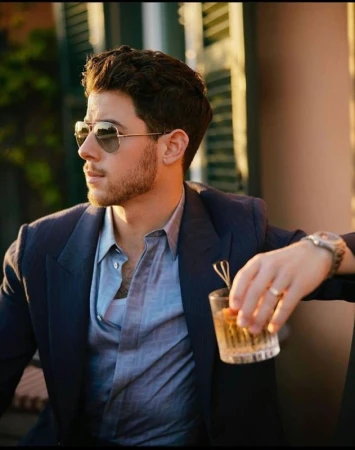 Nick Jonas wearing sunglasses, navy blazer, and blue shirt while holding a glass with hair styled by Charley Mcewen.