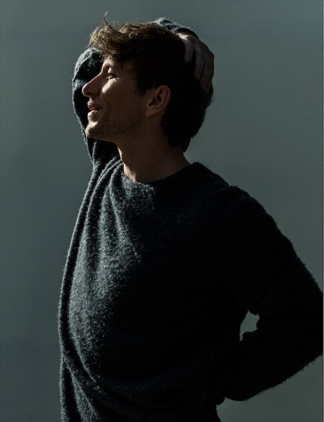 Profile portrait of adult male in sweater with hand on head. Soft, diffused light highlights the side of his face against a gray background.