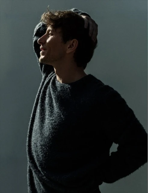 Profile portrait of adult male in sweater with hand on head. Soft, diffused light highlights the side of his face against a gray background.
