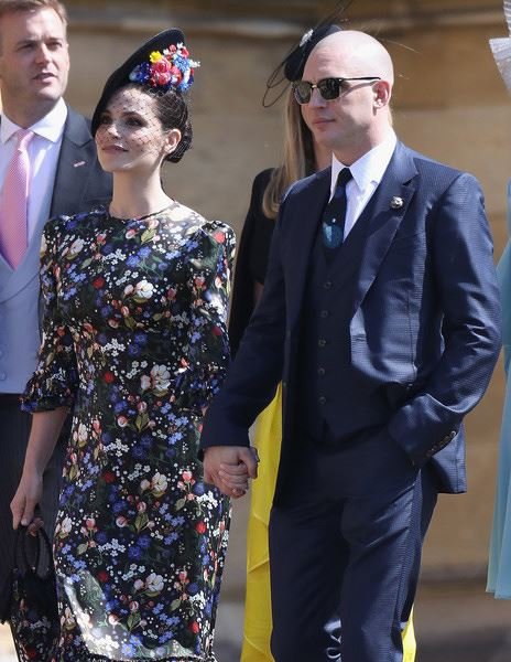 Two people holding hands. Woman in floral dress and fascinator, man in dark suit and sunglasses. Event in daylight, possible wedding.