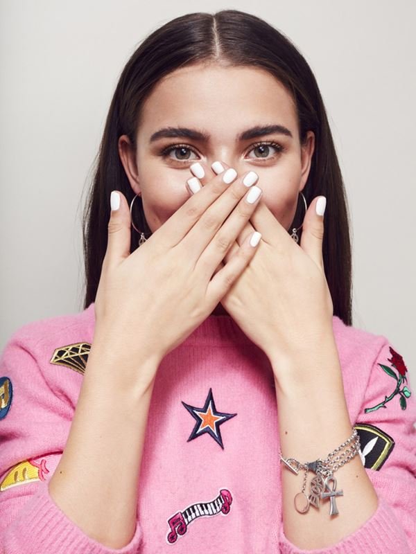 Portrait of a smiling young woman with brunette hair, wearing a pink sweater with patches, covering her mouth with white manicured hands.