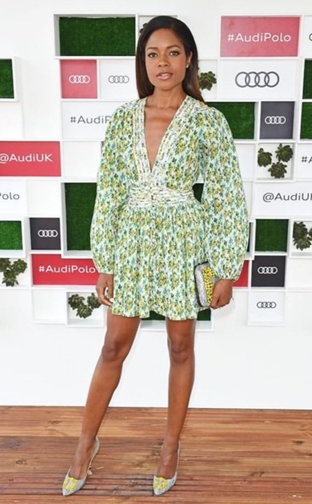 Adult female poses in short floral dress and heels at outdoor promotional event with branded logos on modular background.