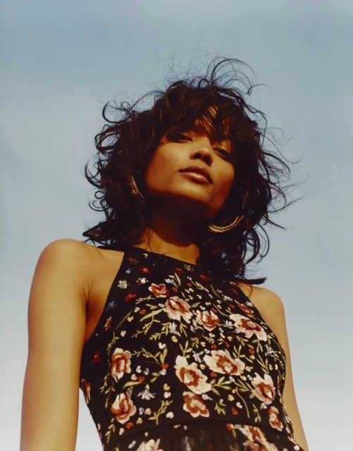 Brunette woman with short, curly hair, wearing a floral embroidered top, gazes up against a clear, blue sky in a low-angle portrait.
