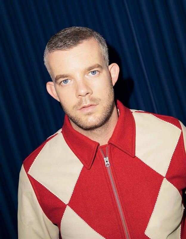 Portrait of a white adult male with short grey hair, wearing a red and beige patterned jacket in front of a blue curtain.