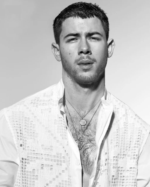 Black and white portrait of an adult male wearing a patterned white shirt with layers of necklaces in a neutral studio setting.