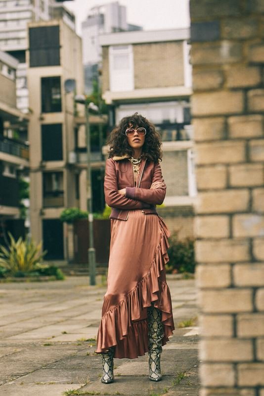 Woman in mauve jacket and satin skirt standing in an urban courtyard with apartment buildings in the background on an overcast day.