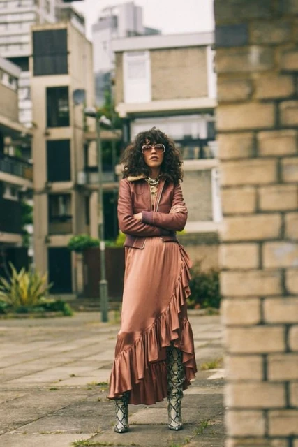 Woman in mauve jacket and satin skirt standing in an urban courtyard with apartment buildings in the background on an overcast day.