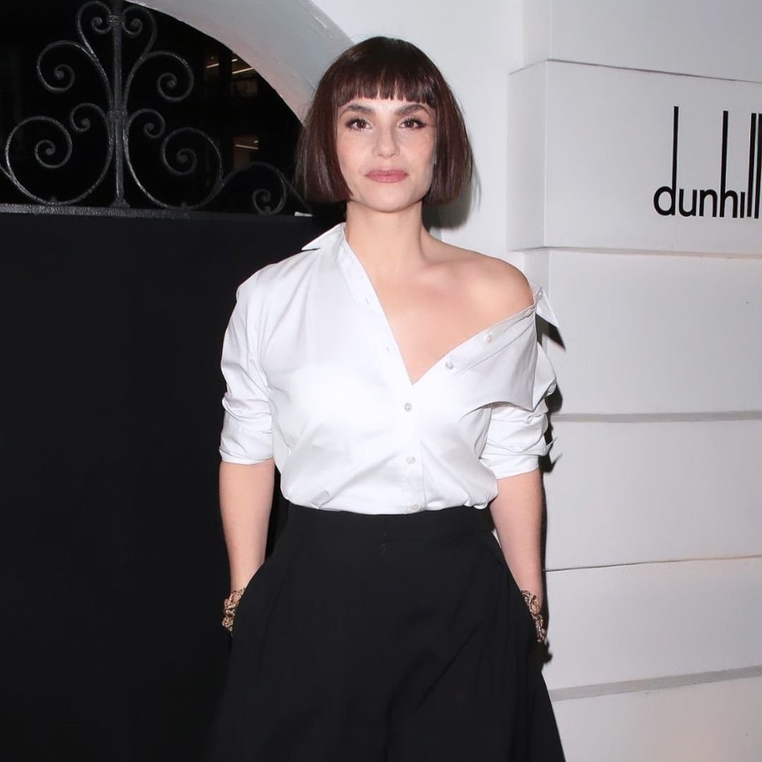 A brunette woman with bangs poses in a white, off-the-shoulder shirt and black pants against a branded, neutral-toned backdrop.