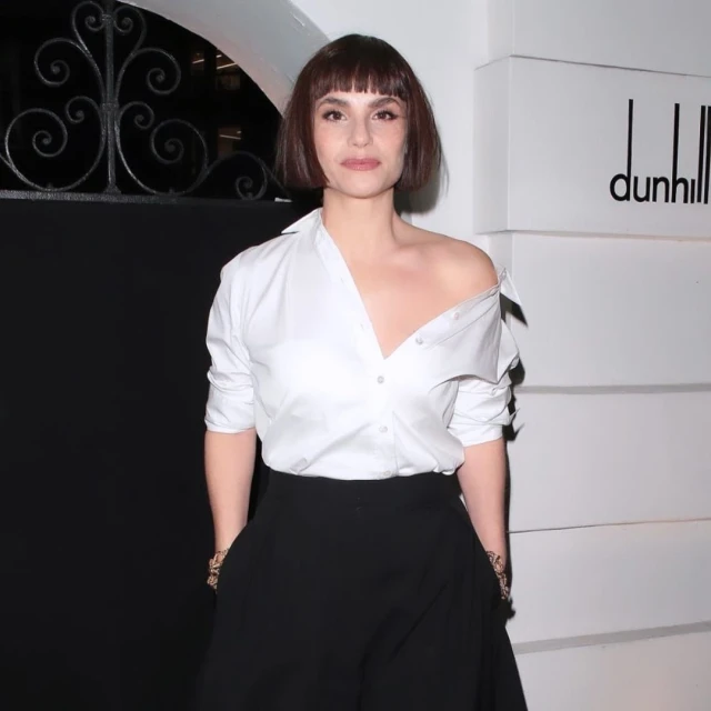 A brunette woman with bangs poses in a white, off-the-shoulder shirt and black pants against a branded, neutral-toned backdrop.