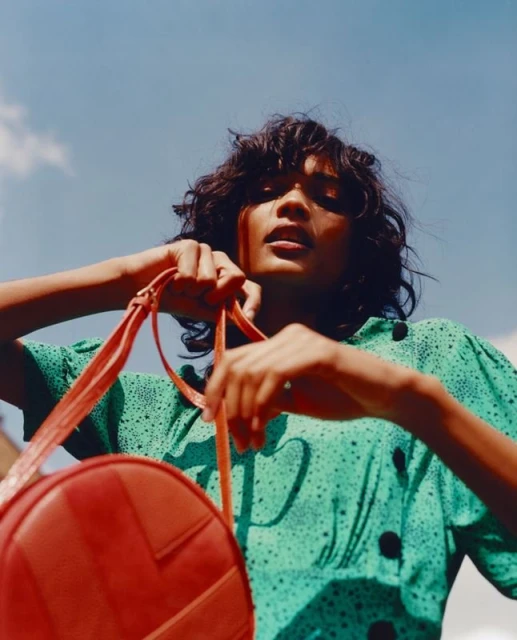 Fashionable woman in stylish green polka dot blouse and red circle bag, blue sky background, adds a vibrant, chic touch.