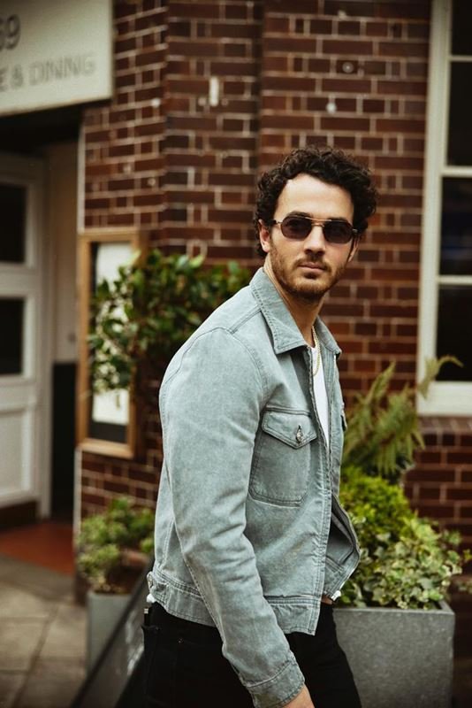 Man wearing a light denim jacket and dark sunglasses posing outdoors in front of a red brick building with planters.