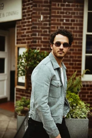 Man wearing a light denim jacket and dark sunglasses posing outdoors in front of a red brick building with planters.