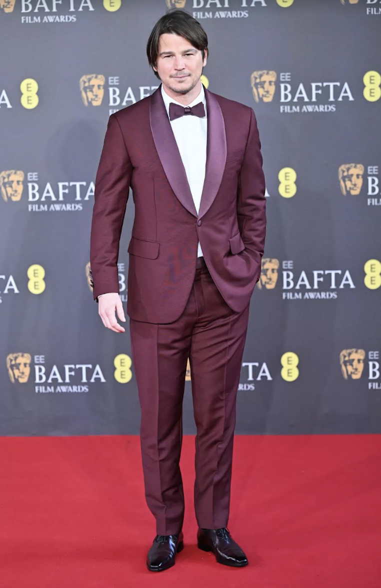 Man in a burgundy suit and bow tie stands on a red carpet in front of a BAFTA Film Awards backdrop at an indoor event.