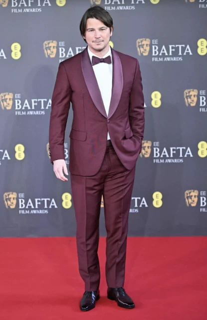 Man in a burgundy suit and bow tie stands on a red carpet in front of a BAFTA Film Awards backdrop at an indoor event.