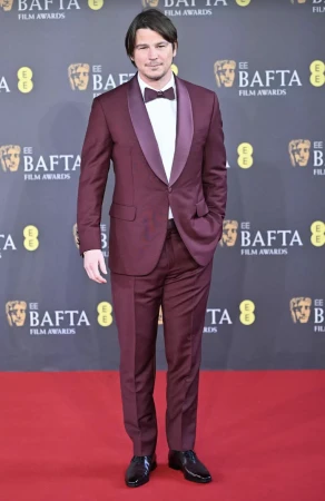 Man in a burgundy suit and bow tie stands on a red carpet in front of a BAFTA Film Awards backdrop at an indoor event.