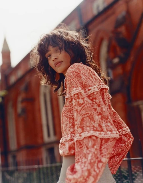 A brunette woman poses in front of a red brick building, wearing a patterned blouse, captured in natural lighting with a low angle.