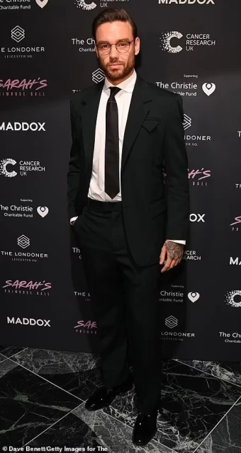 A man in a dark suit and glasses stands in front of a backdrop featuring event logos, providing a formal and posed portrait.