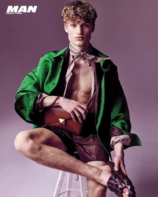 A young adult male sits on a stool in a studio, wearing a green shirt, brown shorts, and sandals, holding a brown bag. 