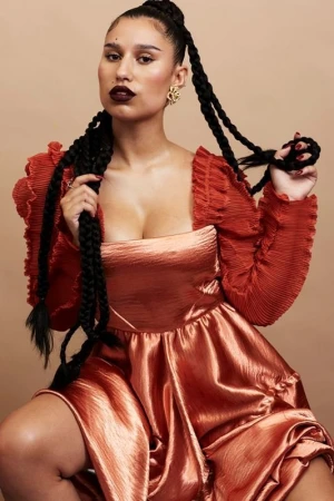 Portrait of a woman with dark lipstick and long braided hair, wearing a shiny copper-colored dress with pleated sleeves, seated indoors.