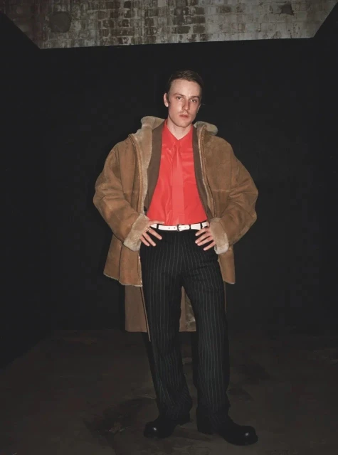 A fair-skinned man in a tan shearling coat and pinstripe pants stands indoors against a dark background, hands on hips, looking at the camera.