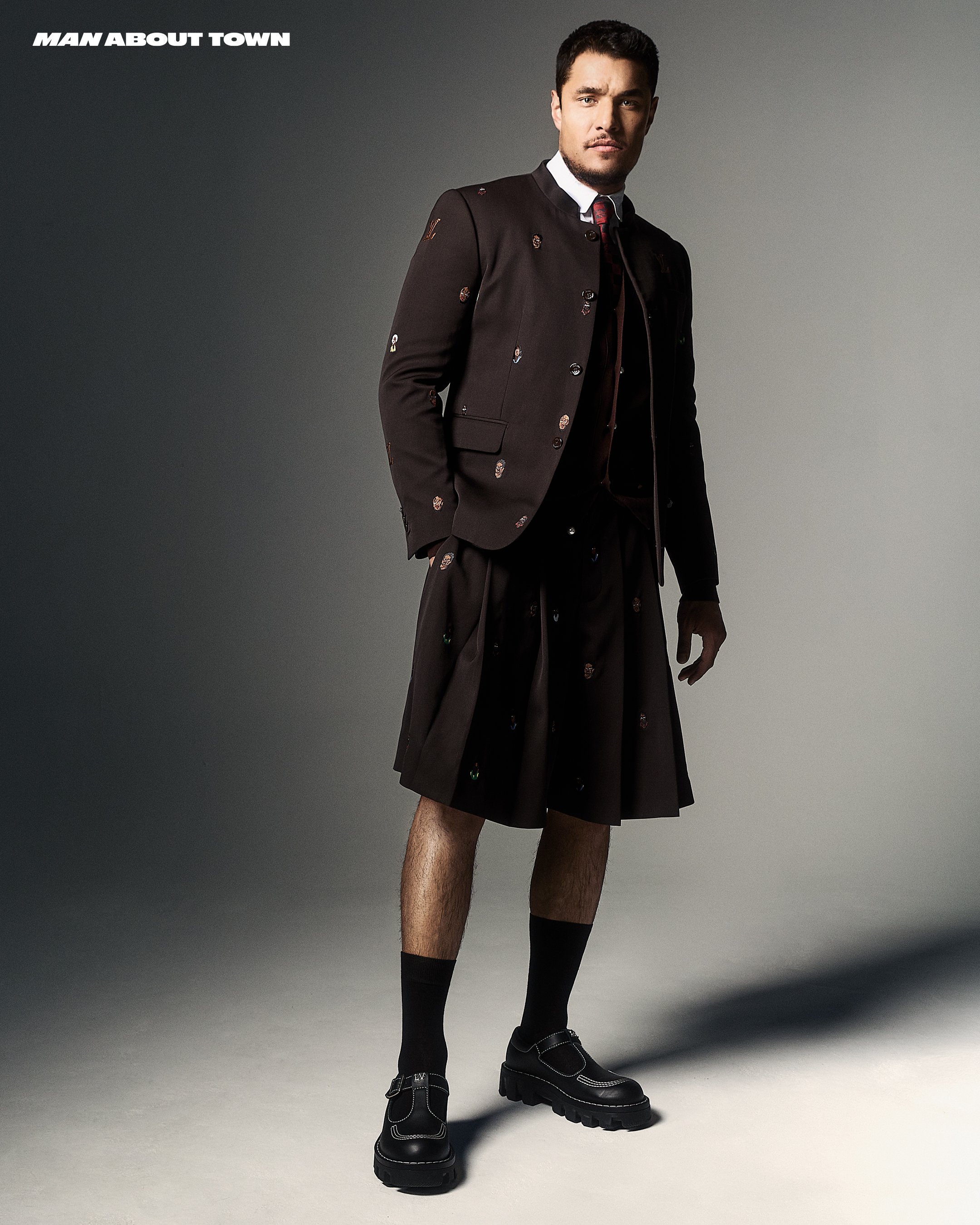 Fashion shot of a man in a dark kilt suit, posing in a photo studio with bright light and gray background for 'Man About Town'.