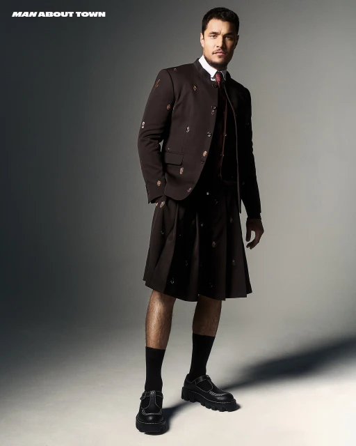Fashion shot of a man in a dark kilt suit, posing in a photo studio with bright light and gray background for 'Man About Town'.